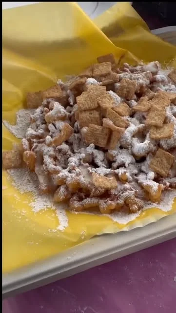 A funnel cake with powdered sugar, cereal pieces, and a sweet drizzle